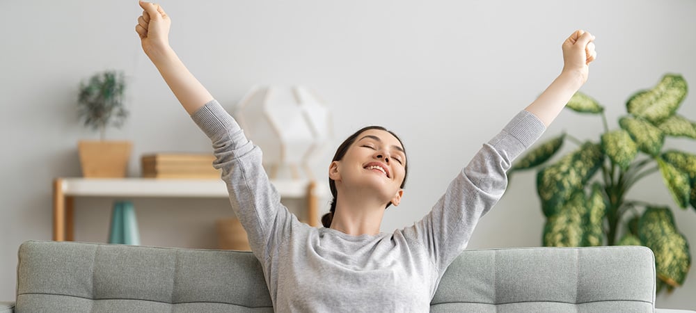 Happy woman on her sofa at home stretching arms. cbd tincture online. tincture for sleep. sleep tincture. tincture cbd oil.